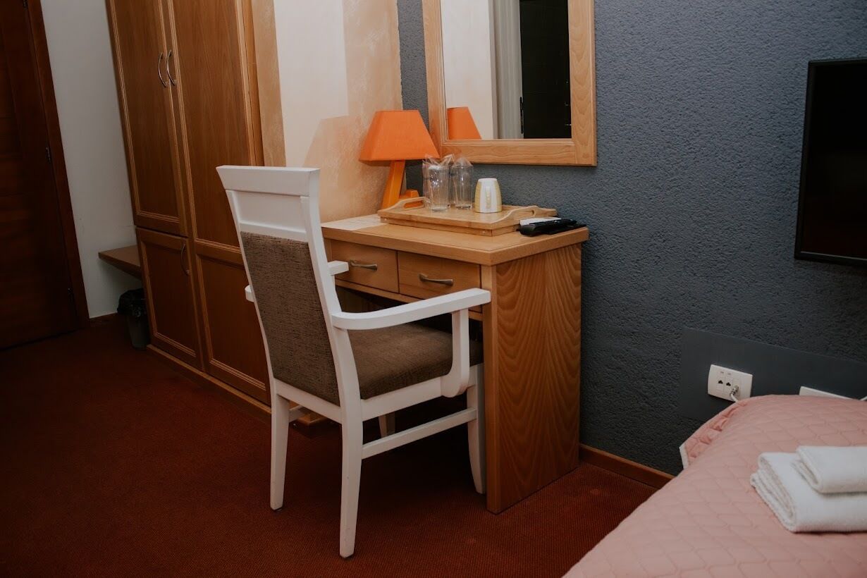 A hotel room with a wooden desk, a chair, a bed with a pink quilt, a mirror, and a television on the wall.