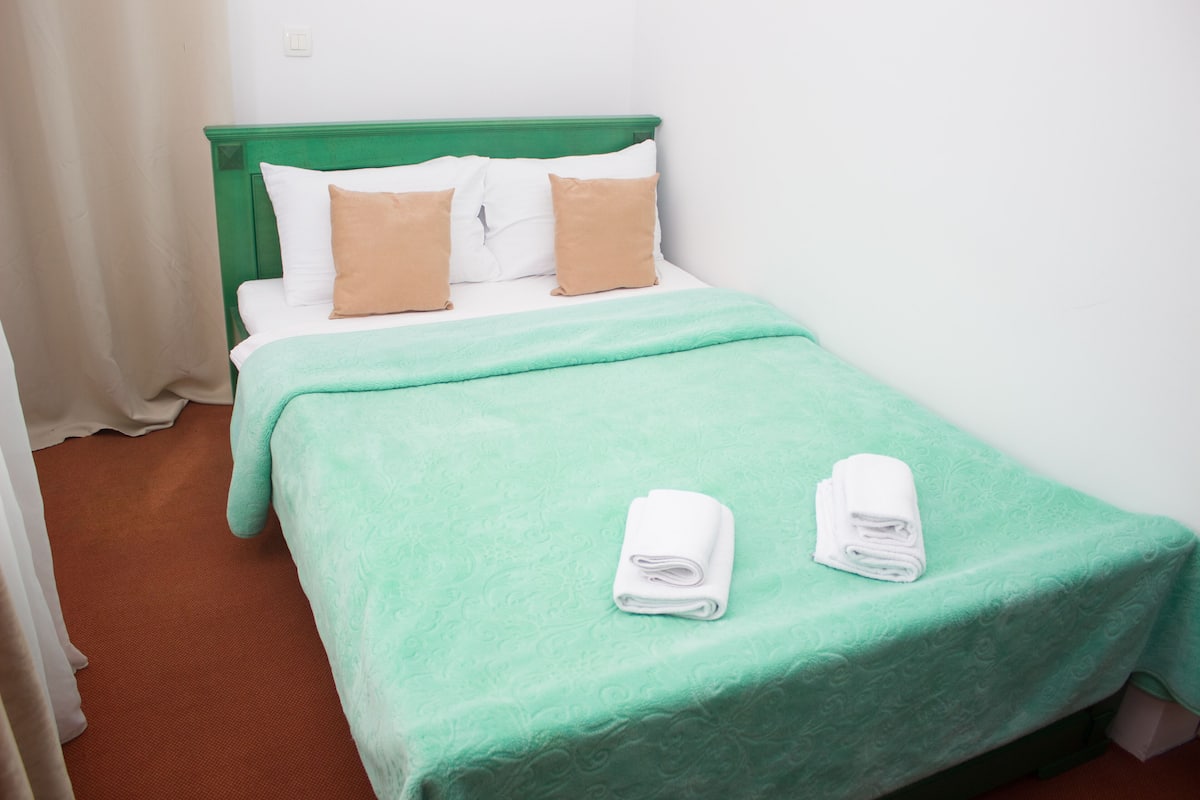 Uma cama de solteiro com cabeceira verde, travesseiros brancos e castanhos, e um edredom verde. Há duas toalhas brancas dobradas sobre a cama.
