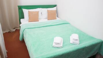 Una cama individual con cabecera verde, almohadas blancas y marrones, y colcha verde. Hay dos toallas blancas enrolladas sobre la cama.