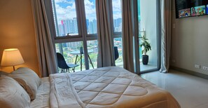 Habitación de hotel con una cama grande, una silla, una mesa y vistas a la ciudad a través de amplias ventanas.