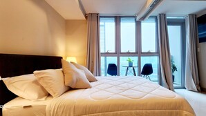 A hotel room with a large bed, a headboard, a window with curtains, a small table with a plant, and a chair.