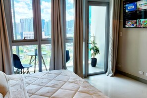 Habitación de hotel con una cama grande, una mesita pequeña, dos sillas, una planta en maceta y vistas a la ciudad a través de la ventana.