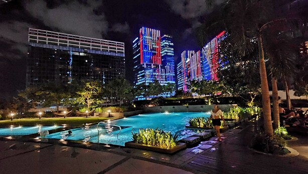 A nighttime scene of a swimming pool area with illuminated buildings in the background.