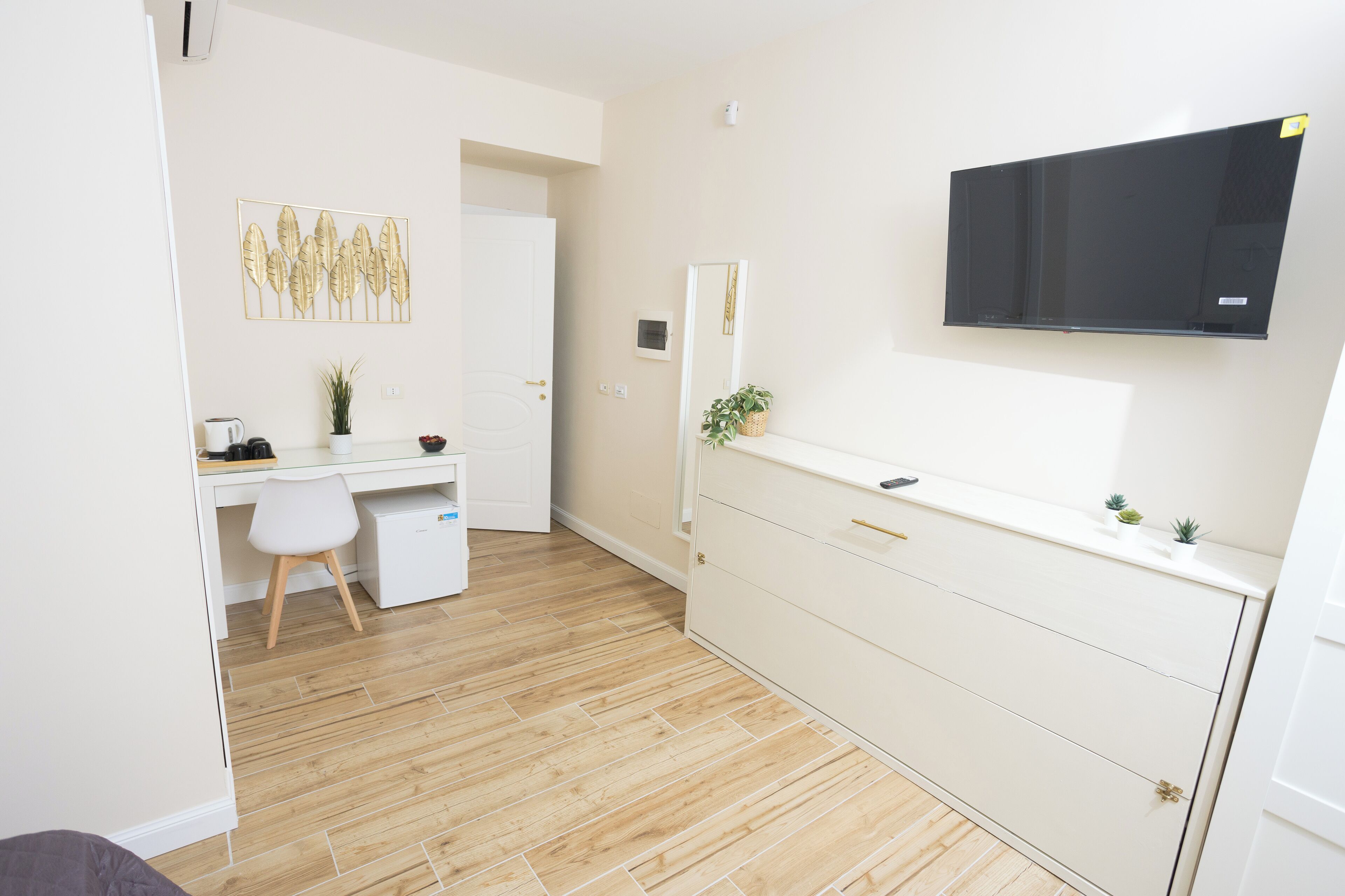 Una habitación moderna y minimalista con piso de madera, un escritorio blanco con silla, un armario blanco, un televisor montado y una pequeña planta en la cómoda.