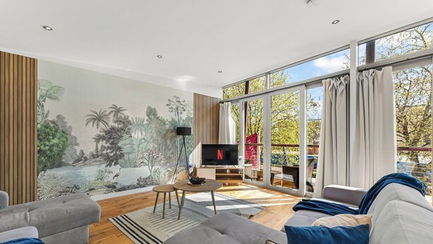 A modern living room with a large TV, grey sofas, a mural, and a view of trees through large windows. - Modern Luxe Living in Lincoln - Flat 3 (Lincolnshire)