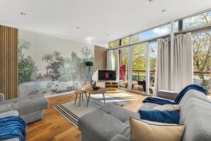 A modern living room with a large TV, grey sofas, a mural, and a view of trees through large windows. - Modern Luxe Living in Lincoln - Flat 3 (Lincolnshire)