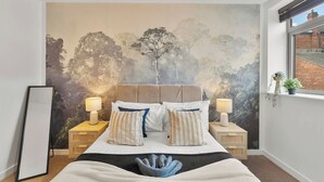 A bedroom with a large bed, two bedside tables with lamps, a wall mural, a window, and a view of a brick building. - Modern Luxe Living in Lincoln - Flat 3 (Lincolnshire)
