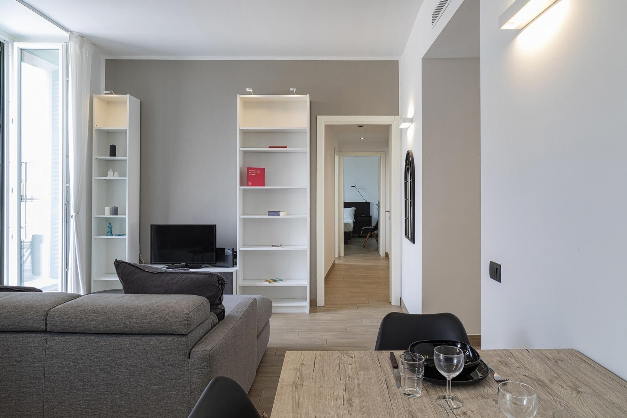 A modern living room with a grey sofa, a TV, and a dining area with a table set for two.