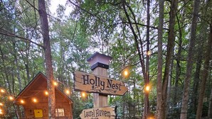 Un cartel de madera con indicaciones para 'Holly Nest' y 'Laurel Haven' en un área boscosa, con una cabaña al fondo.