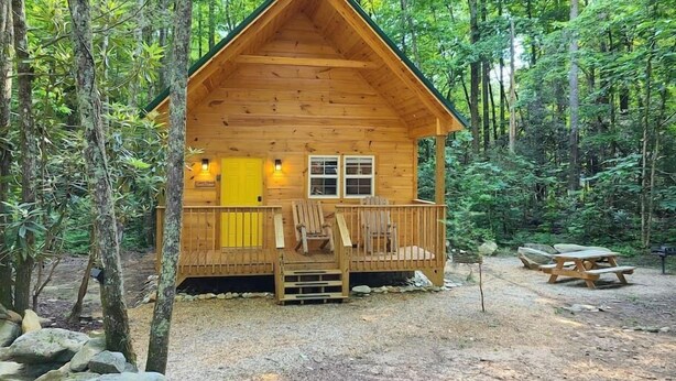 Una cabaña de madera con porche, una puerta amarilla y una mesa de picnic en un entorno boscoso.