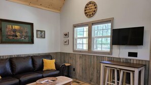 Una sala de estar con techo de madera, un sofá de cuero negro, una mesa de centro de madera, un televisor montado en la pared y una ventana con vistas a árboles.