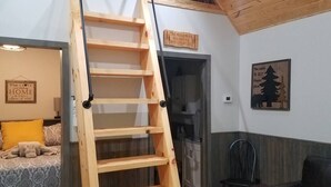 Ein Zimmer mit einer Holzleiter, die zu einem Schlafbereich führt, einem Bett mit gelbem Kissen, einem schwarzen Stuhl und einem Holzschild an der Wand.