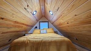 Ein gemütliches Schlafzimmer im Dachgeschoss mit schrägen Wänden, einem Bett mit gelber Bettdecke und einem Dachfenster darüber.