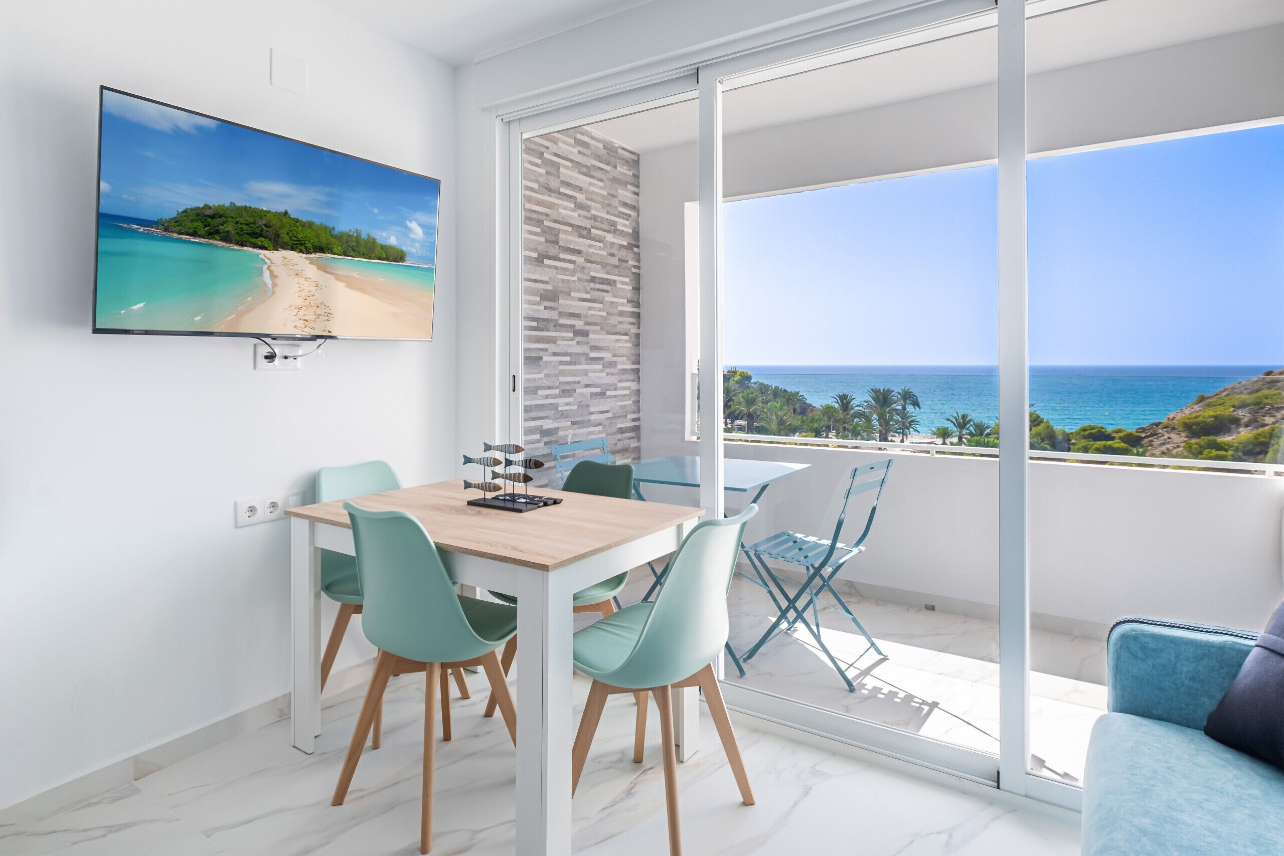 Un salon moderne comprenant un coin repas, un téléviseur fixé au mur et un balcon avec vue sur la mer.