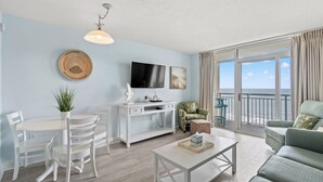 A modern living room with a white dining set, a white coffee table, a sofa, a TV, and a balcony with a view of the sea. - Bay Watch Resort 1239 -Oceanfront-Water Amenities (North Myrtle Beach)