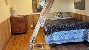 Un dormitorio tipo loft con una escalera de madera que lleva a una zona de descanso elevada, un aparador de madera, una colcha estampada y un espejo en la pared.