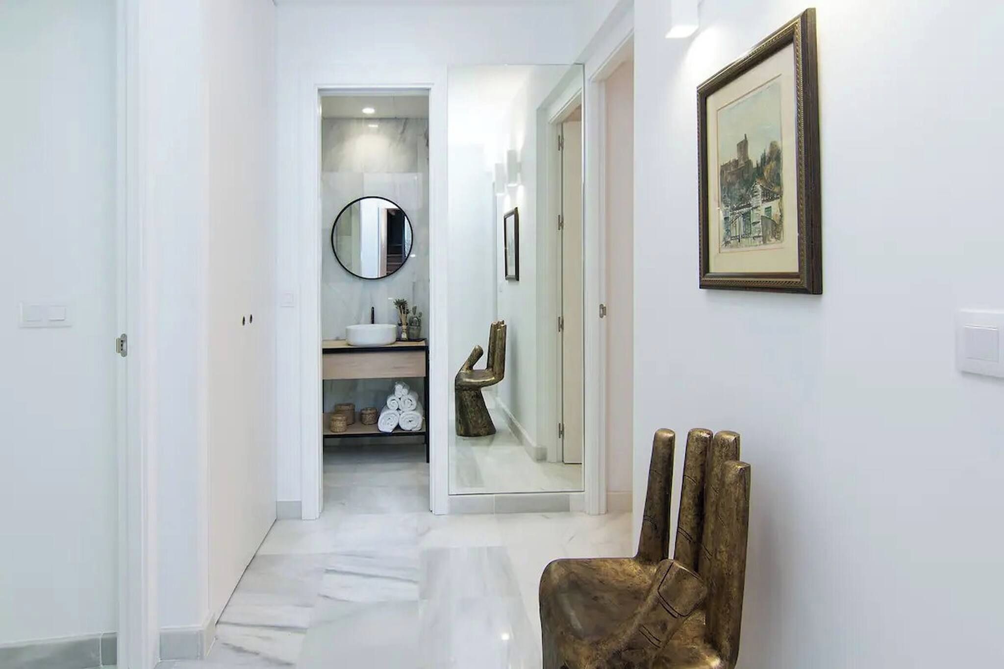 Un pasillo con piso blanco, un espejo redondo, un cuadro enmarcado en la pared y una silla.