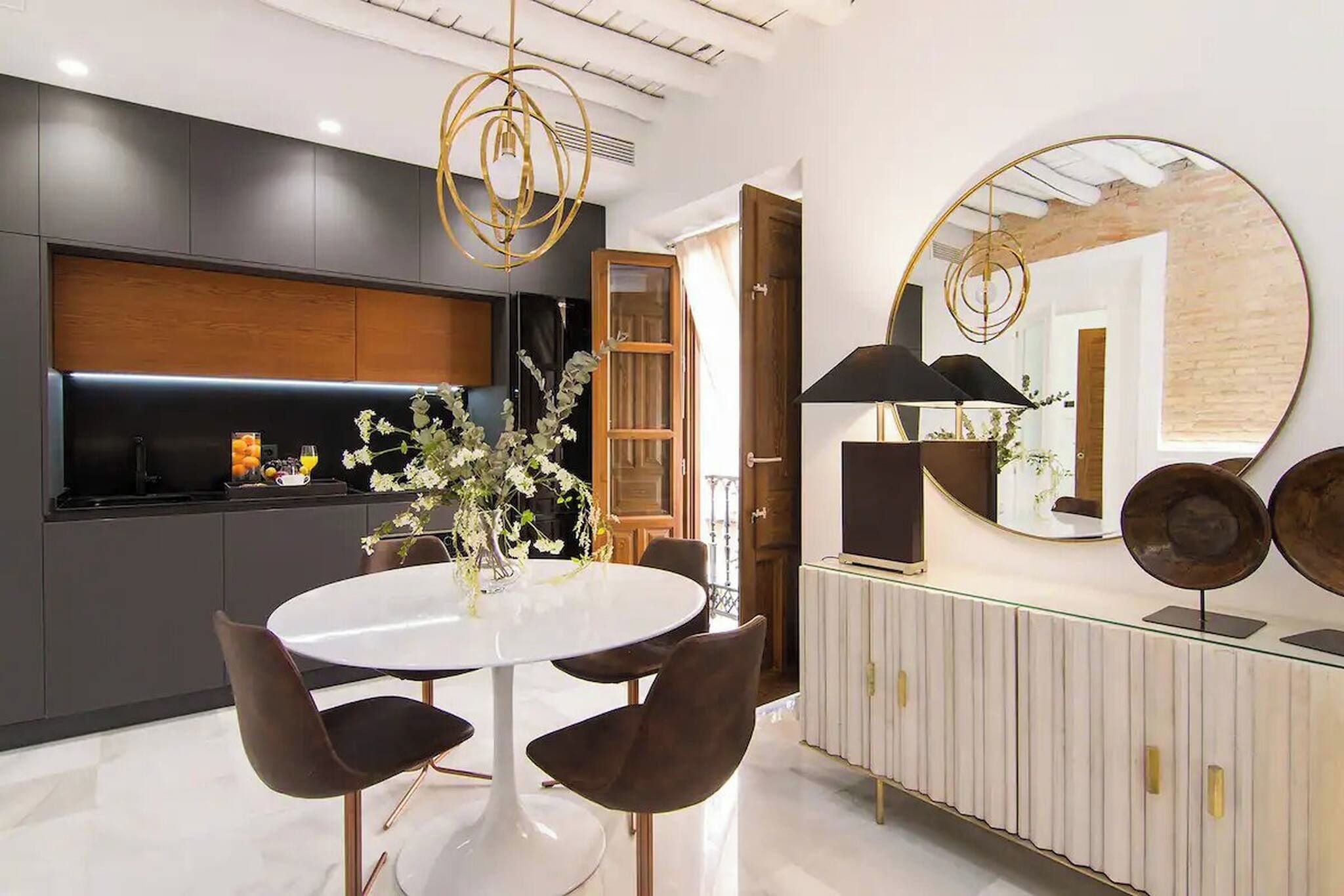 Una cocina moderna con una isla central, un comedor con una mesa redonda y sillas, y un espejo grande en la pared.