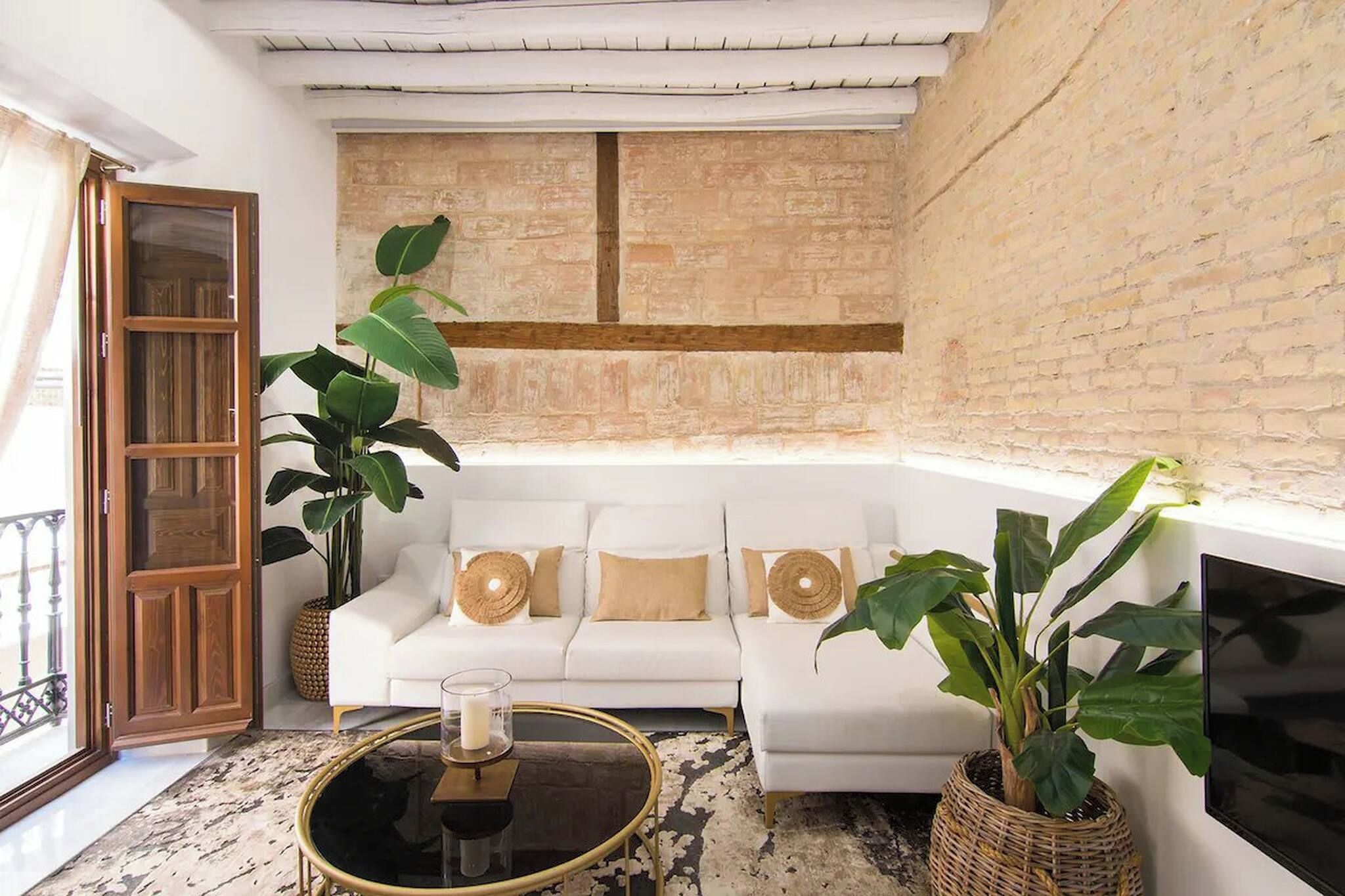 Una sala de estar con un sofá blanco, dos plantas verdes grandes, una mesa de centro redonda con una vela, y un televisor montado en la pared.