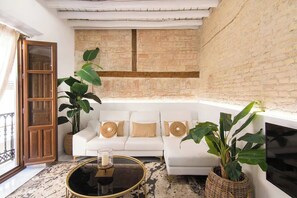 A living room with a white sofa, two large green plants, a round coffee table with a candle, and a TV mounted on the wall. - Riad Alhambra 2 (granada)