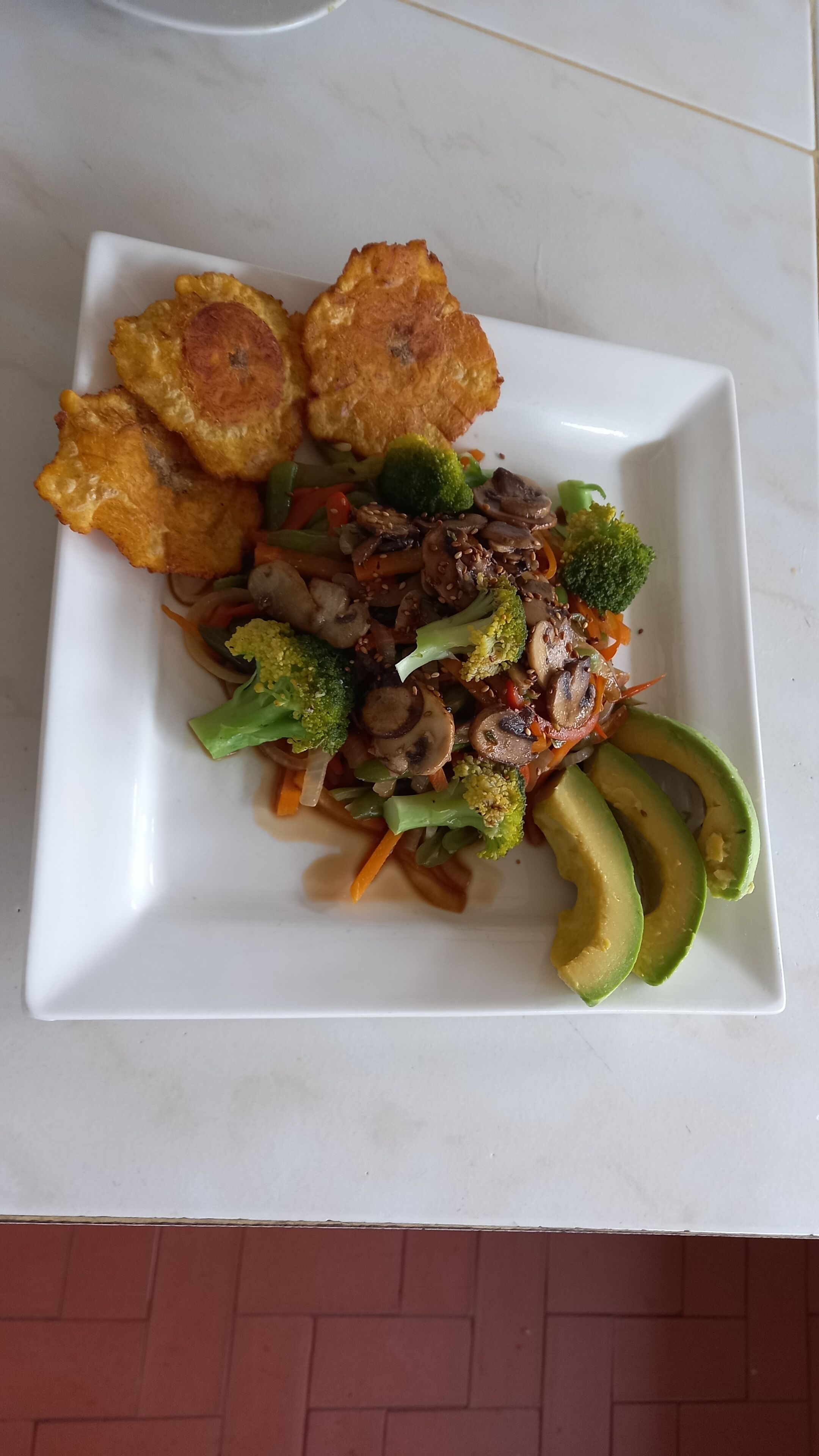 Un plato cuadrado blanco con un salteado que incluye brócoli, champiñones en rodajas, zanahorias y rebanadas de aguacate, acompañado de dos alimentos fritos.