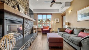 A cozy living room with a stone fireplace, a large window, a ceiling fan, and a grey sofa with red cushions. - Wild Horses | Pool | Hot Tub | Hot Springs (Radium Hot Springs)