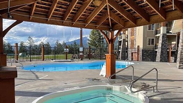 A swimming pool with a wooden deck overhead, surrounded by a fenced area and a building with stone pillars. - Wild Horses | Pool | Hot Tub | Hot Springs (Radium Hot Springs)