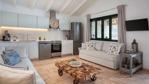 Una cocina moderna con armarios blancos, horno empotrado y lavavajillas. Hay una mesa de centro de madera con un cuenco encima, un sofá blanco con cojines estampados y una pequeña mesita con una linterna.