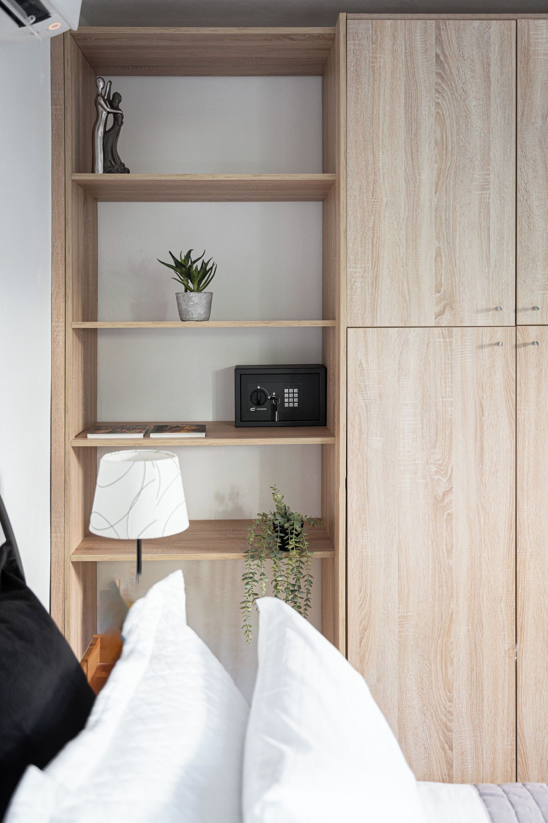 A bedroom with a bed, a wooden wardrobe, a shelf with a plant, a lamp, and a framed picture.