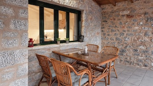 Una habitación con paredes de piedra y techo de madera, un ventanal con contraventanas verdes y una mesa de comedor con sillas de mimbre.