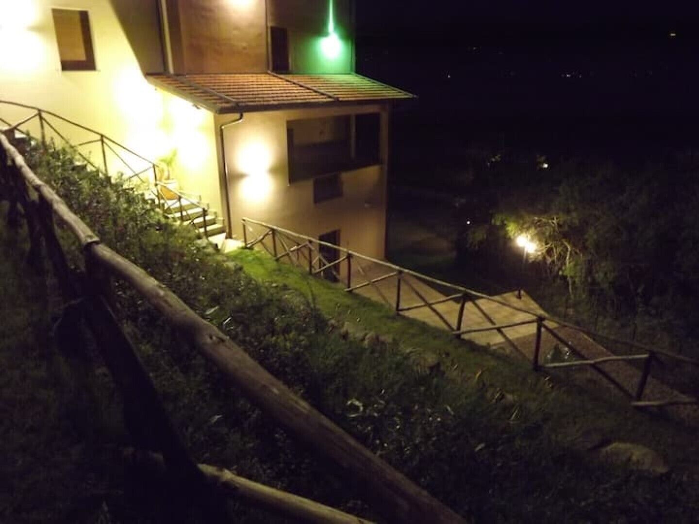 Una escalera conduce a un edificio con una torre iluminada en verde, rodeado de vegetación y una cerca de madera.