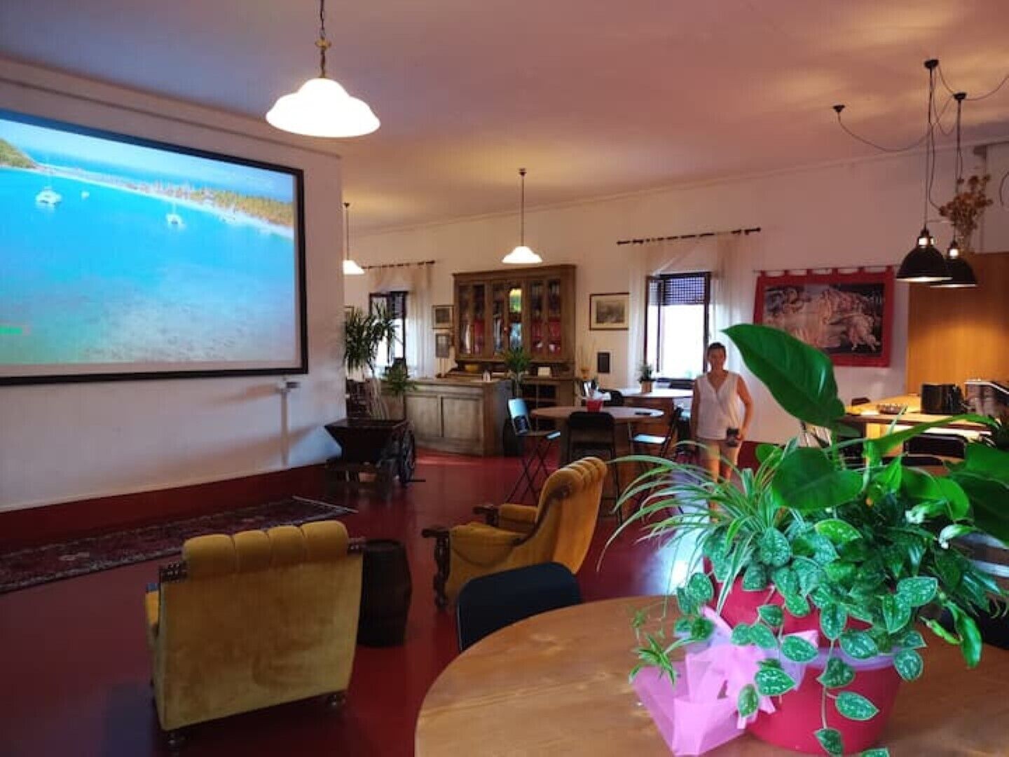 Eine großzügige Lobby mit einem großen Bildschirm, der einen malerischen Ausblick zeigt, bequemen Sitzgelegenheiten, einem Essbereich und einer Küche im Hintergrund.