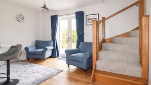 A living room with a wooden staircase, two blue armchairs, a grey armchair, a white table, and a clock on the wall. - Chynoweth House (Newquay)