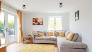 A living room with a grey sectional sofa, a yellow rug, a wooden coffee table, and a lamp. There is a painting on the wall and a window with curtains. - Chynoweth House (Newquay)