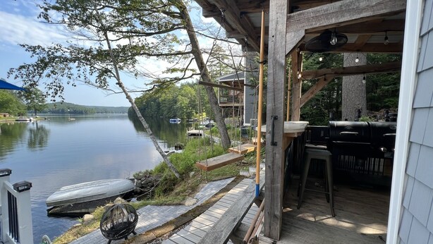A wooden deck with a grill and outdoor seating, overlooking a calm lake with boats and docked cars. - Lake Front Retreat with Deep Water Dock (Weare)