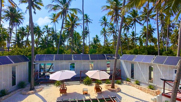 A resort with multiple rooms, each with a balcony and solar panels on the roof. There are two umbrellas and a few chairs in the courtyard. - Zanzibar Beach House- West (Dongwe)