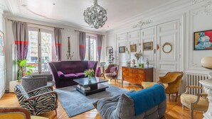 A spacious living room with a large purple sofa, a grey sofa, and a wooden coffee table. There is a chandelier hanging from the ceiling, and a fireplace with a mantel. - Sophisticated and Grand Apt. in Perfect Location! (Paris)