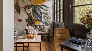 Un comedor con una mesa dispuesta para dos, un sofá, un mural floral y una ventana con vistas a árboles.