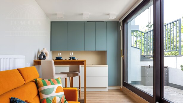 A modern living room with a sofa, a coffee table with books and fruit, and a view of a kitchen and balcony. - ArtSquare by Fragola Apartments (Kraków)