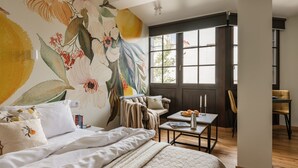 A bedroom with a large floral mural, a bed with white bedding, a bedside table with books, and a view of the outside through a window. - ArtSquare by Fragola Apartments (Kraków)