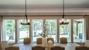 Un coin repas avec une table et des chaises en bois, de grandes fenêtres donnant sur la verdure et des lampes suspendues.