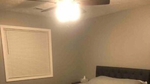 Une chambre à coucher avec un lit, une table de chevet, un ventilateur de plafond et une fenêtre avec des stores.