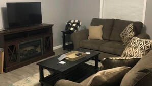 Un salon comprenant un canapé, une table basse, un téléviseur posé sur un meuble en bois et une fenêtre avec des stores.