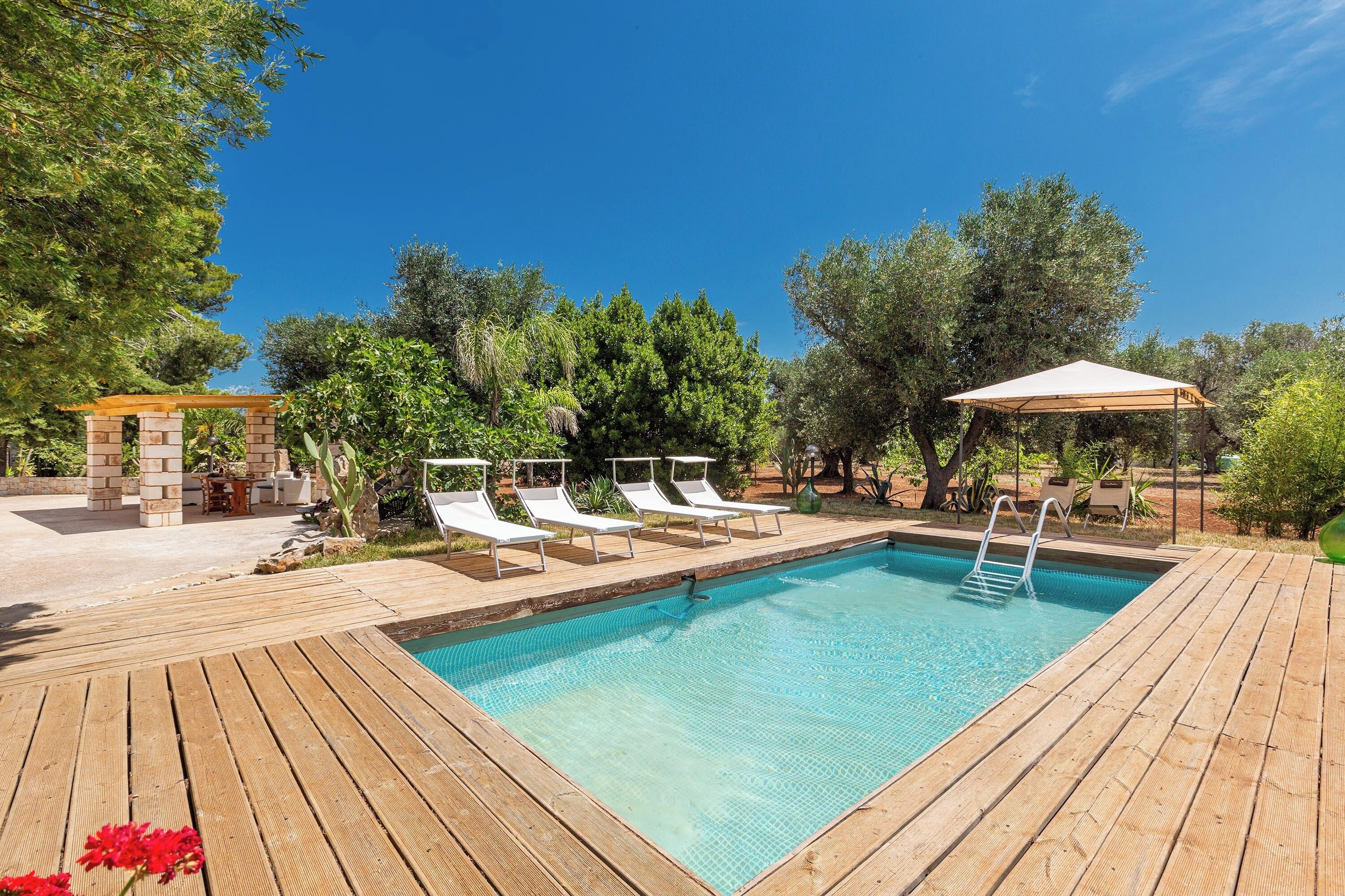 A rectangular swimming pool with a wooden deck, surrounded by lounge chairs and a pergola.