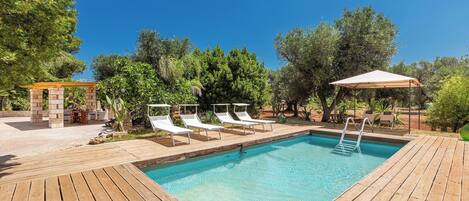 A rectangular swimming pool with a wooden deck, surrounded by lounge chairs and a pergola.
