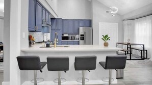 A modern kitchen with blue cabinets, a central island with bar stools, and a dining area with a table and chairs. - Luxury & Bright ~ 2 Master BR ~ Games ~ Backyard! (Houston)