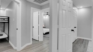 A hallway with white walls and a wooden floor, leading to a bedroom with a bed and a nightstand. - Luxury & Bright ~ 2 Master BR ~ Games ~ Backyard! (Houston)