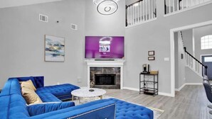 A modern living room with a blue sectional sofa, a fireplace, a flat-screen TV, and a staircase leading to an upper level. - Luxury & Bright ~ 2 Master BR ~ Games ~ Backyard! (Houston)