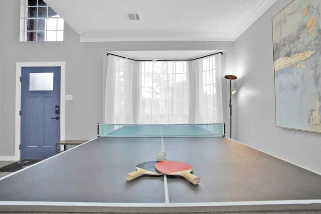 Une table de ping-pong dans une pièce avec une porte bleue, une grande peinture abstraite et un lampadaire.