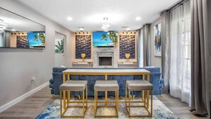 A modern living room with a fireplace, a blue sofa, and three bar stools. - Luxury Home 4 Rooms ~ Pool Table & Poker~ King Bed (Houston)
